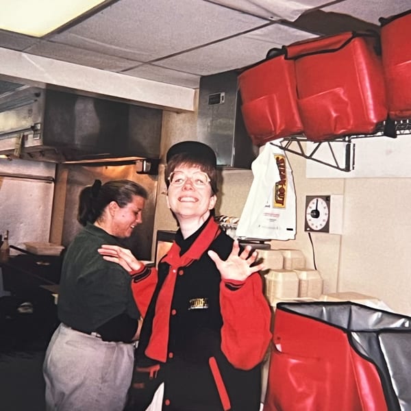 Two women in a restaurant kitchen, one smiling and gesturing, with red delivery bags overhead.