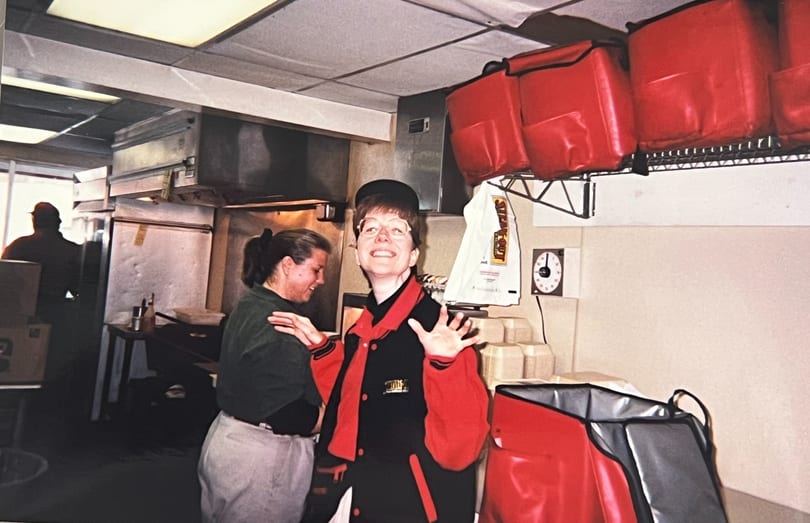 Two women in a restaurant kitchen, one smiling and gesturing, with red delivery bags overhead.