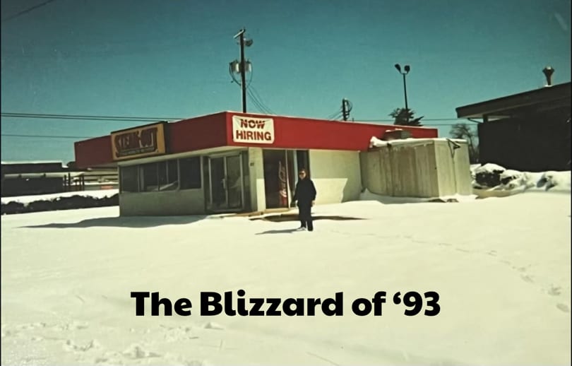 A person stands in deep snow outside a Steak-Out restaurant with a "Now Hiring" sign.