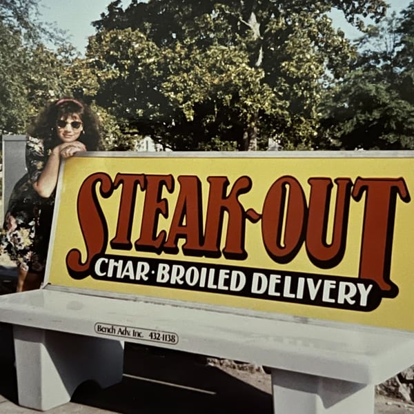 Woman in sunglasses leaning on a "Steak-Out Char-Broiled Delivery" sign on a bench.