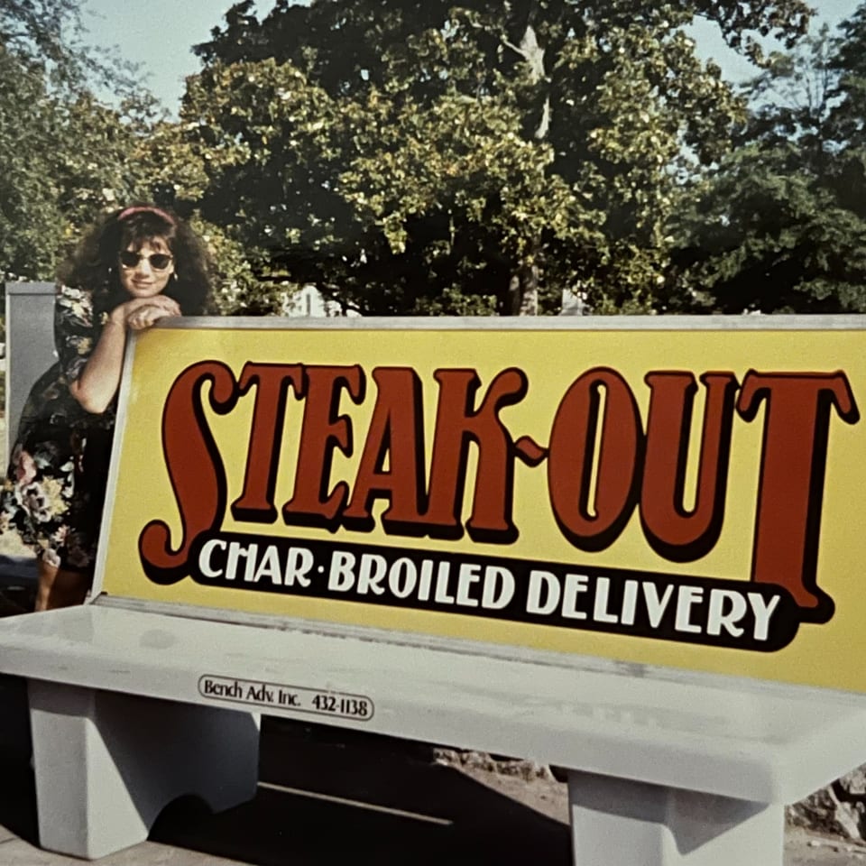 Woman in sunglasses leaning on a "Steak-Out Char-Broiled Delivery" sign on a bench.