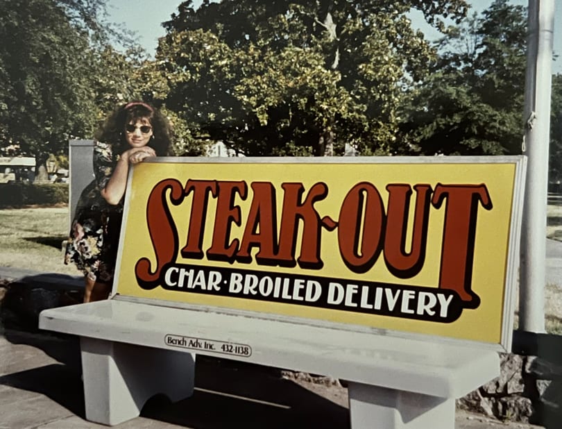 Woman in sunglasses leaning on a "Steak-Out Char-Broiled Delivery" sign on a bench.