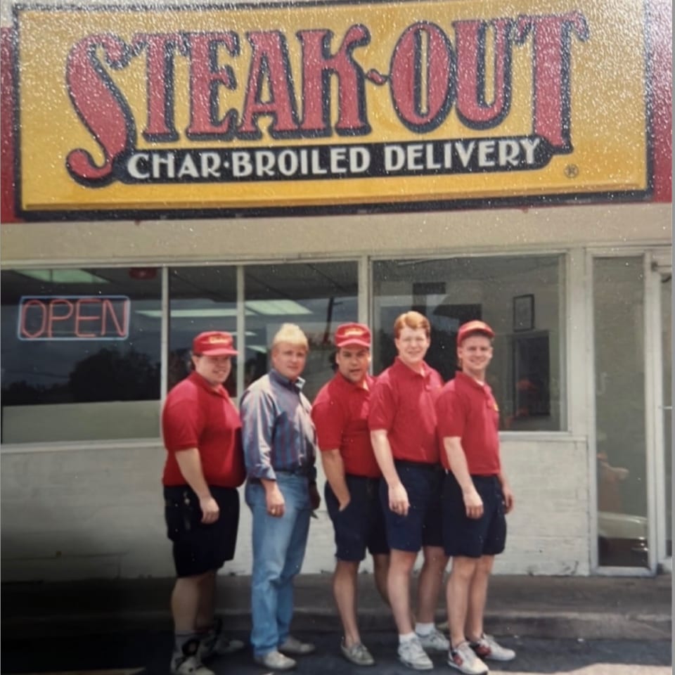 Five young men, four in red uniforms, stand outside a Steak-Out Char-Broiled Delivery restaurant.