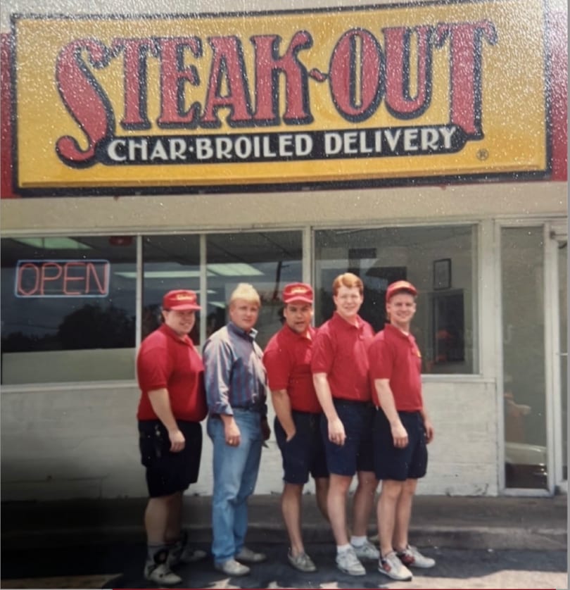 Five young men, four in red uniforms, stand outside a Steak-Out Char-Broiled Delivery restaurant.